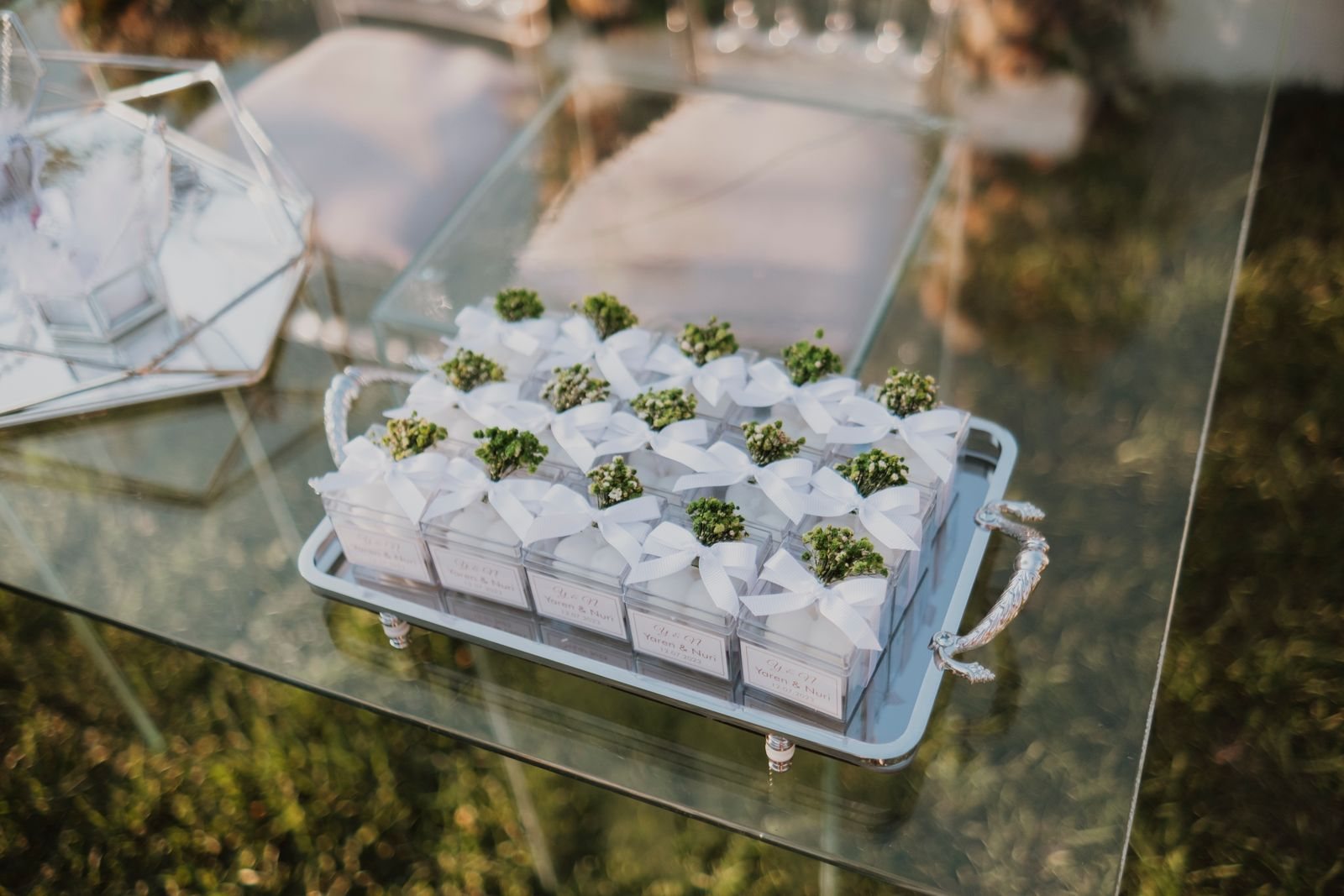 Witte bedankjes met strikjes en groene takjes op een glazen tafel buiten bij een bruiloft
