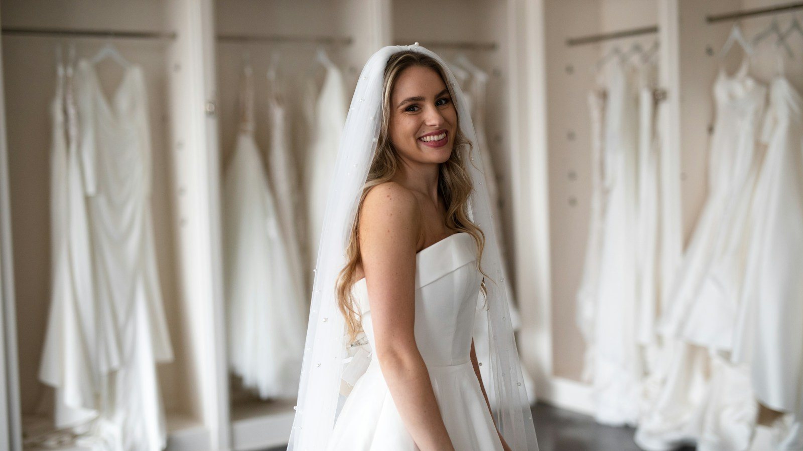 Lachende bruid met sluier in een strapless trouwjurk staat in een bruidsmodezaak voor rekken vol witte trouwjurken