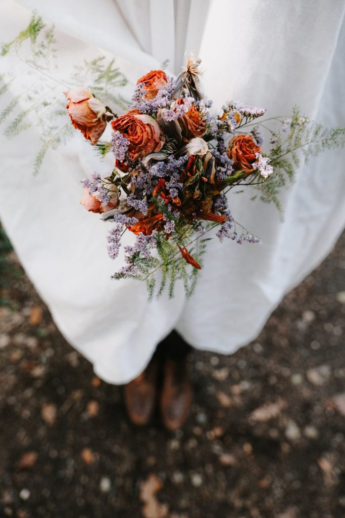 Bruid houdt een rustiek bruidsboeket met roestoranje rozen en gedroogde bloemen boven herfstbladeren