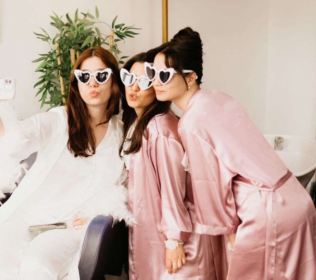 Bruid en bruidsmeisjes in roze badjassen met hartjesbrillen maken samen een vrolijke foto