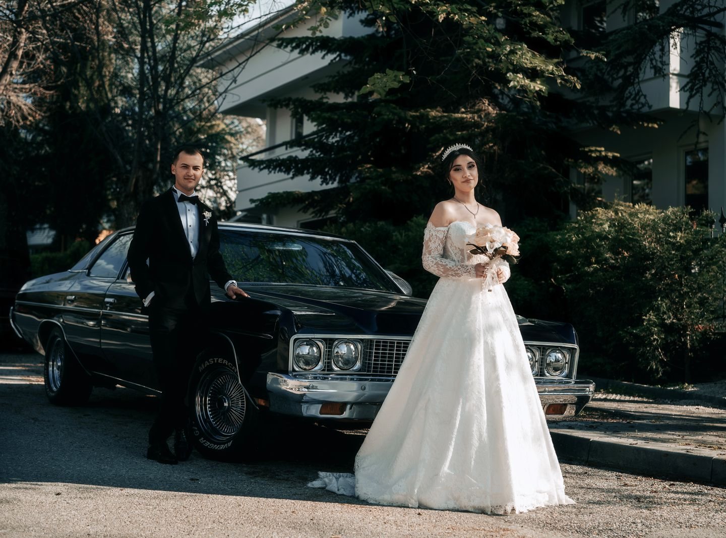 Bruidspaar in trouwjurk en smoking poseert naast een donkere klassieke Amerikaanse oldtimer