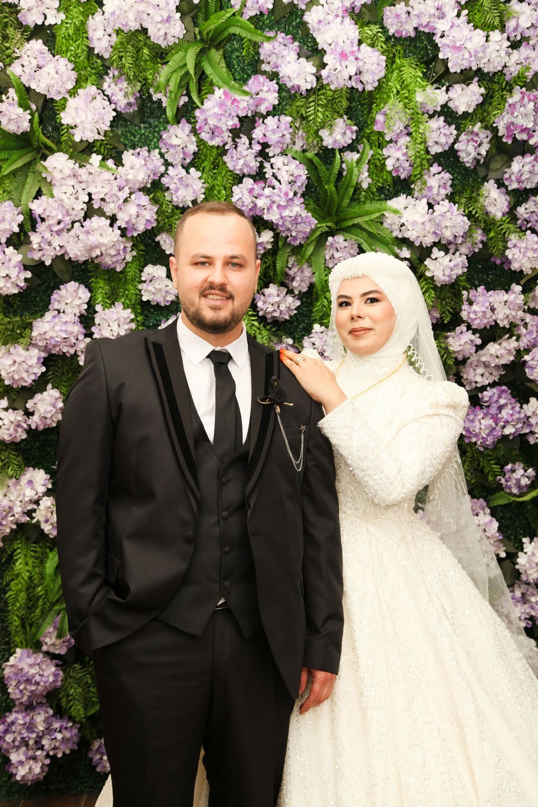 Bruidspaar poseert lachend voor een weelderige bloemenwand in roze en paars