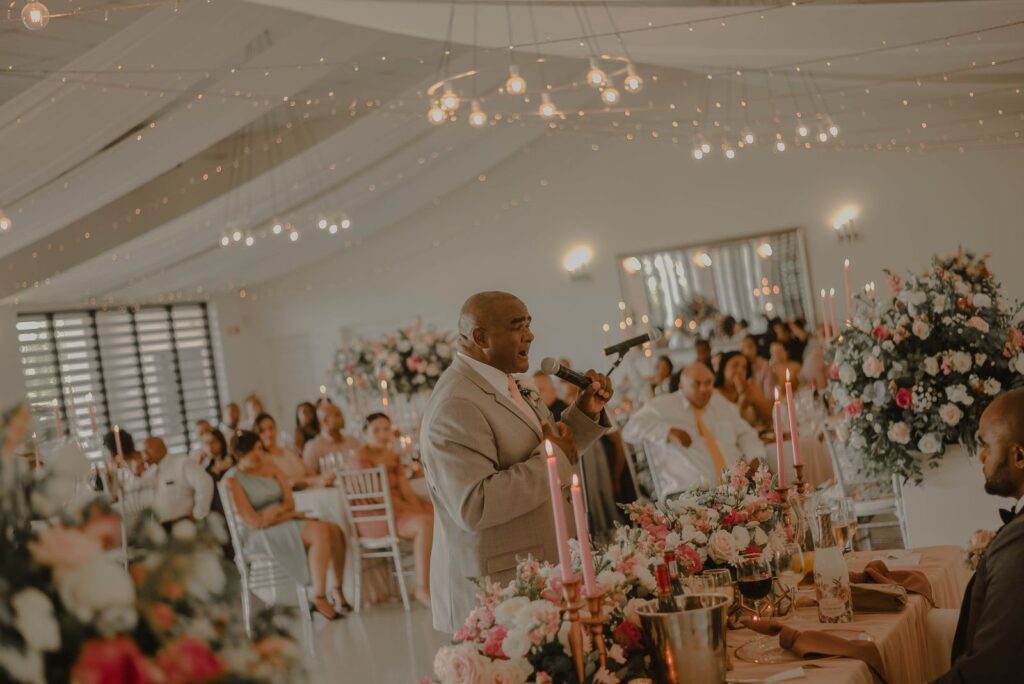 Man houdt een speech met een microfoon tijdens het diner, met gasten aan feestelijk gedekte tafels vol bloemen