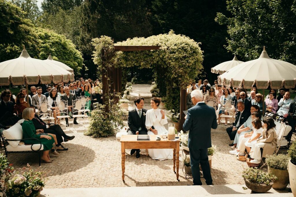 Bruidspaar zit aan een houten tafel in een tuin terwijl de trouwambtenaar spreekt, omringd door gasten onder parasols