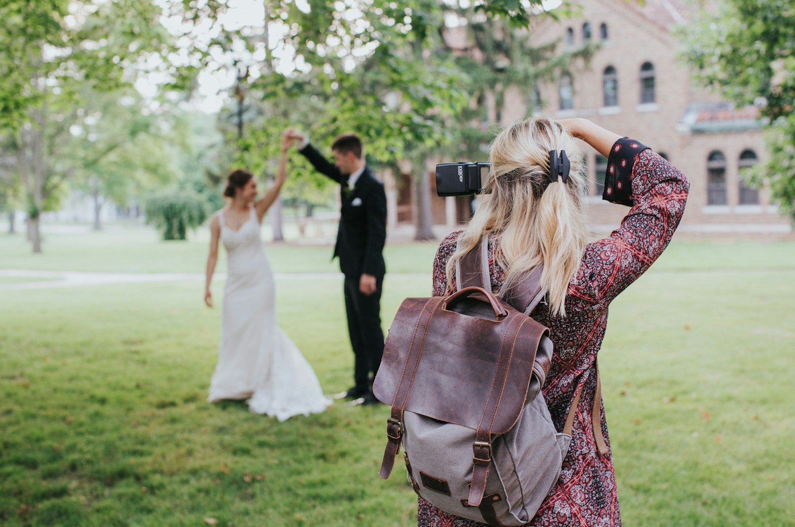 Trouwfotografe met camera en rugzak fotografeert een lachend bruidspaar dat danst op het gras in een park